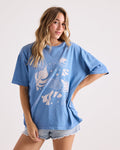 A woman with long wavy hair wears the Volcom Women's Stone Breaker Tee—a loose-fitting, 100% cotton shirt featuring blue fabric with white graphic designs—paired with light denim shorts. She stands against a plain light background, one hand resting on her hip.