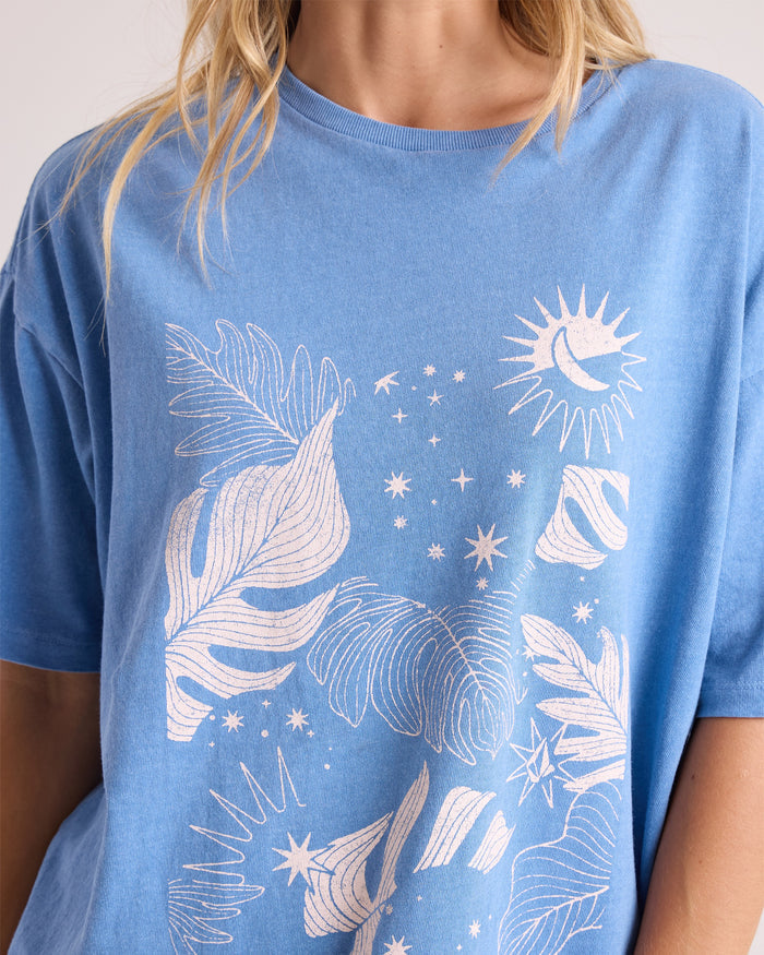 A person is shown from the upper body wearing the Volcom Women's Stone Breaker Tee in blue, a loose-fit 100% cotton shirt featuring white line art with leaves, stars, and a crescent moon on the front.