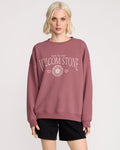 A woman with short blonde hair wears a mauve Volcom Women's Stone Crew and black shorts, standing against a plain light background. The sweatshirt features "TRUE TO THIS VOLCOM STONE" and a sun graphic on the front.