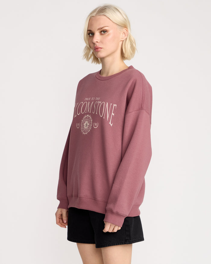 A person with short blonde hair wears the Volcom Women's Stone Crew, a mauve fleece pullover with white text and emblem, paired with black shorts, standing against a plain white background.