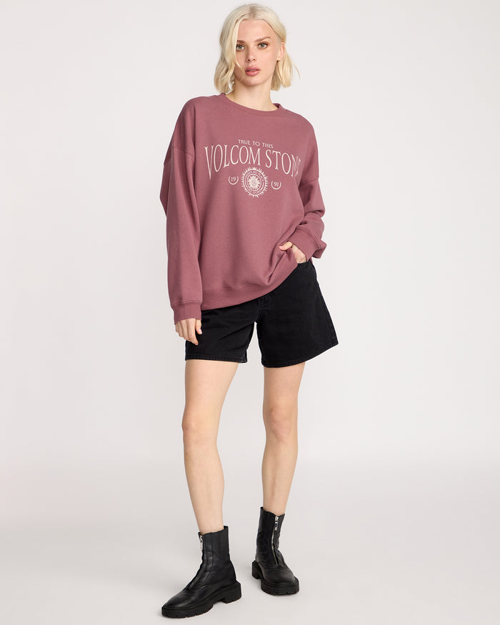 A person with short blonde hair wears a loose mauve Women's Stone Crew by Volcom, paired with black shorts and black boots, standing against a plain white background.