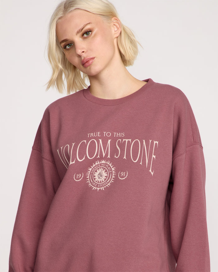 A woman with short blonde hair wears the Volcom Women's Stone Crew, a mauve fleece pullover with "True to this VOLCOM STONE 1991" and a graphic design on the front. She looks slightly to the side with a neutral expression.