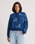 A woman wears the Volcom Women's Stone Dream Hoodie, featuring a blue relaxed fit with a light floral pattern, paired with light blue jeans. She stands against a plain light background, looking at the camera with a neutral expression.