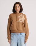 A woman with short brown hair stands facing forward in a Volcom Women's Stone Dream Hoodie—a brown relaxed-fit style featuring a white floral design on the shoulder. She pairs it with light blue jeans, set against a plain light-colored background.