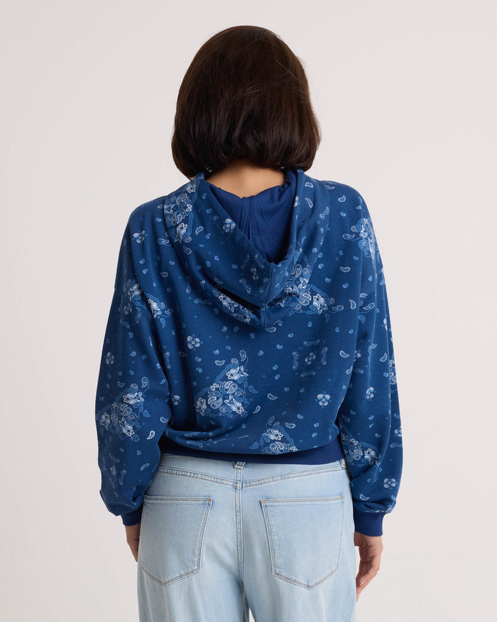 A person with short dark hair is wearing the Volcom Women's Stone Dream Hoodie—a blue patterned relaxed fit hoodie with a kangaroo pocket—and light blue jeans, facing away from the camera against a plain light background.
