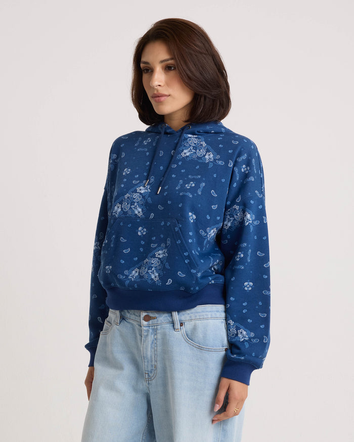 A woman wearing the Volcom Women's Stone Dream Hoodie—a blue, relaxed-fit fleece pullover with a light blue floral pattern and a cozy kangaroo pocket—paired with light blue jeans, stands against a plain light background.