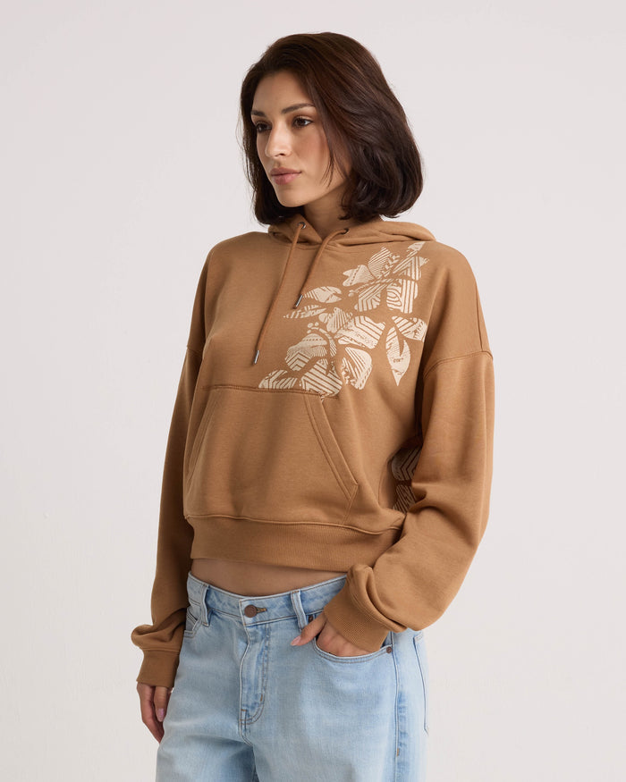 A woman with shoulder-length brown hair wears the Volcom Women's Stone Dream Hoodie—a relaxed-fit style featuring a white leaf design—paired with light blue jeans. She stands against a plain white background with one hand in her pocket.