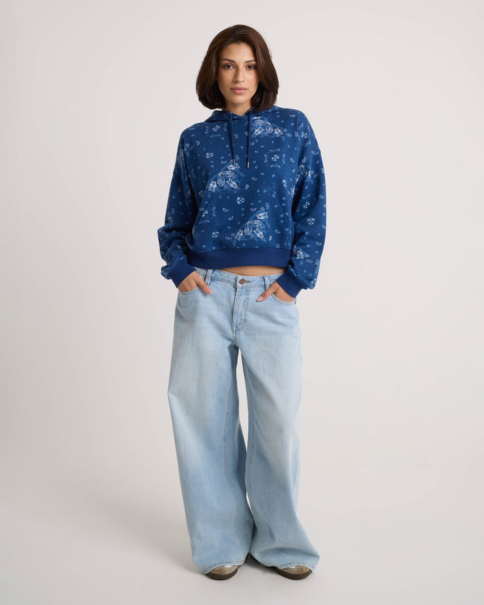 A person stands facing forward, wearing the Volcom Women's Stone Dream Hoodie in a blue floral print with a relaxed fit and kangaroo pocket, paired with light blue wide-leg jeans, hands in pockets, against a plain light background.