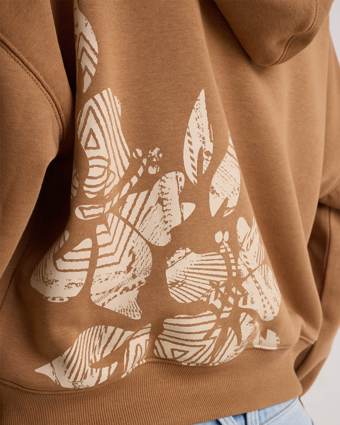 Close-up of the Volcom Women's Stone Dream Hoodie, a relaxed-fit tan hoodie featuring a cream-colored abstract butterfly pattern on the back, styled with blue jeans.
