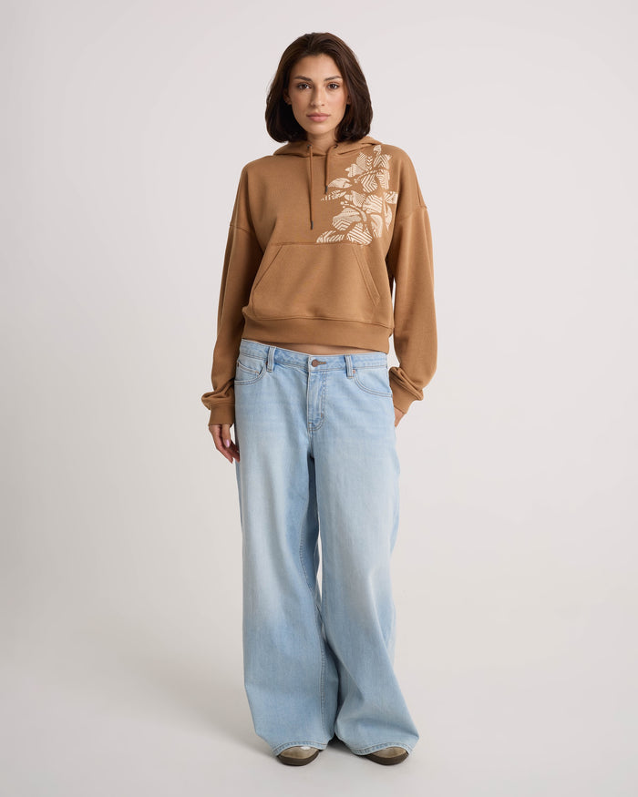 A woman with shoulder-length brown hair and a neutral expression stands against a plain background, wearing the Volcom Women's Stone Dream Hoodie—a relaxed-fit tan fleece pullover with a white floral design—styled with light blue wide-leg jeans.
