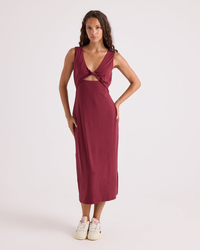 A woman stands against a plain white background wearing the Volcom Women's Stone Luz Dress Solid—a sleeveless, fitted burgundy knit midi dress featuring a front cutout with knot detail—styled with white sneakers.