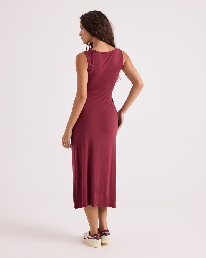 A woman stands facing away against a plain light background, wearing the Volcom Women's Stone Luz Dress Solid in burgundy, styled with cream sneakers featuring burgundy accents.