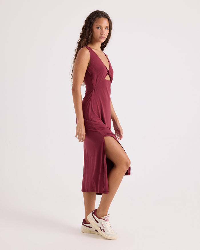 A woman with long curly hair is wearing the Volcom Women's Stone Luz Dress Solid, a fitted maroon midi sleeveless dress featuring a side slit, paired with white sneakers. She stands sideways against a plain white background.