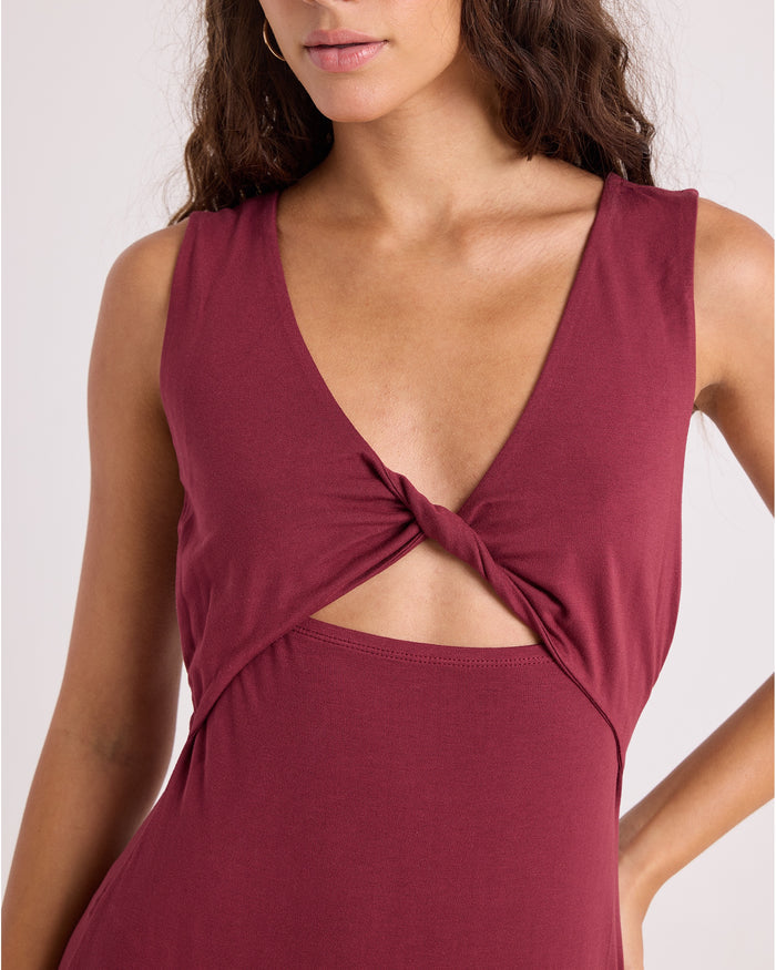 A woman models the Volcom Women's Stone Luz Dress Solid—a fitted, sleeveless maroon dress featuring a knotted front and cutout below the chest—as she poses with one hand on her hip against a light background.