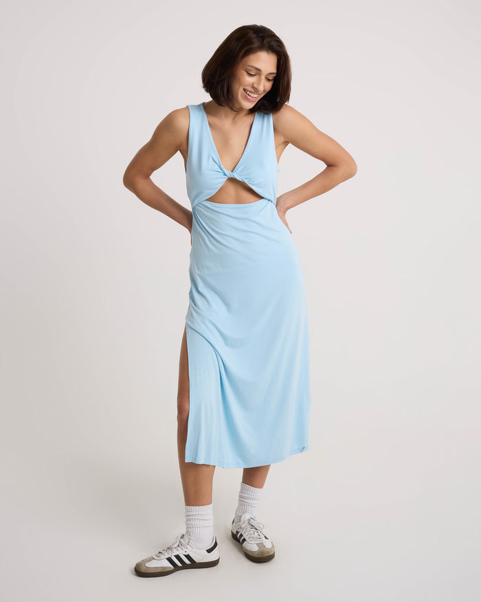 A woman in a Volcom Women's Stone Luz Dress with a twist-front bodice, front cutout, and side slit stands smiling with hands on hips. She has short brown hair, white socks, and white Adidas sneakers against a plain white background.
