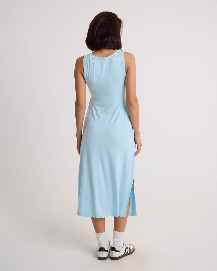 A woman with short dark hair wears the Volcom Women's Stone Luz Dress—a sleeveless midi with a side slit and twist-front bodice—in Cloud Blue, paired with white socks and black-and-white sneakers against a plain light background.