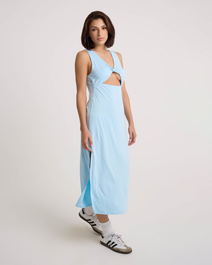 A woman in a Volcom Women's Stone Luz Dress with a twist-front bodice and front ring detail stands against a plain background. She pairs the sleeveless, cut-out midi dress with white socks and black-and-white sneakers.