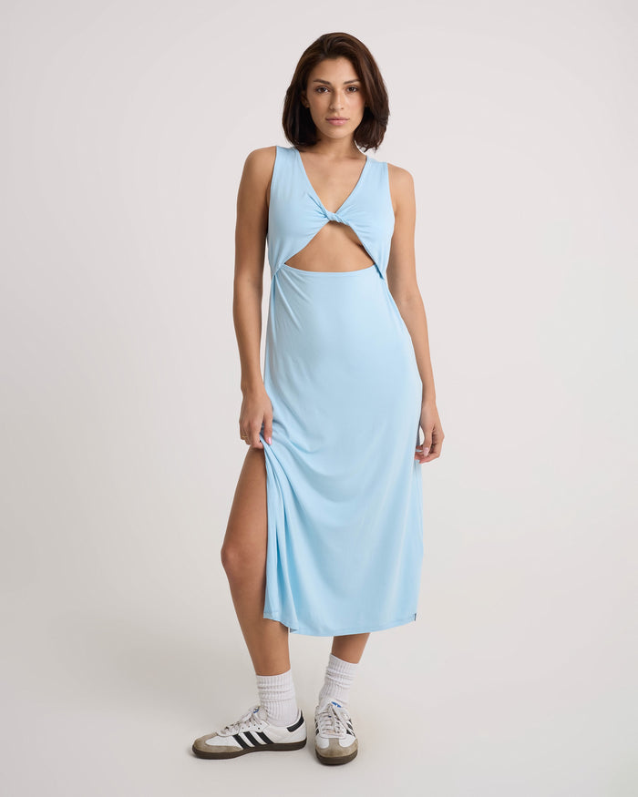 A person wears the Volcom Women's Stone Luz Dress in Cloud Blue, styled with white socks and black-and-white Adidas sneakers, standing against a plain light background.