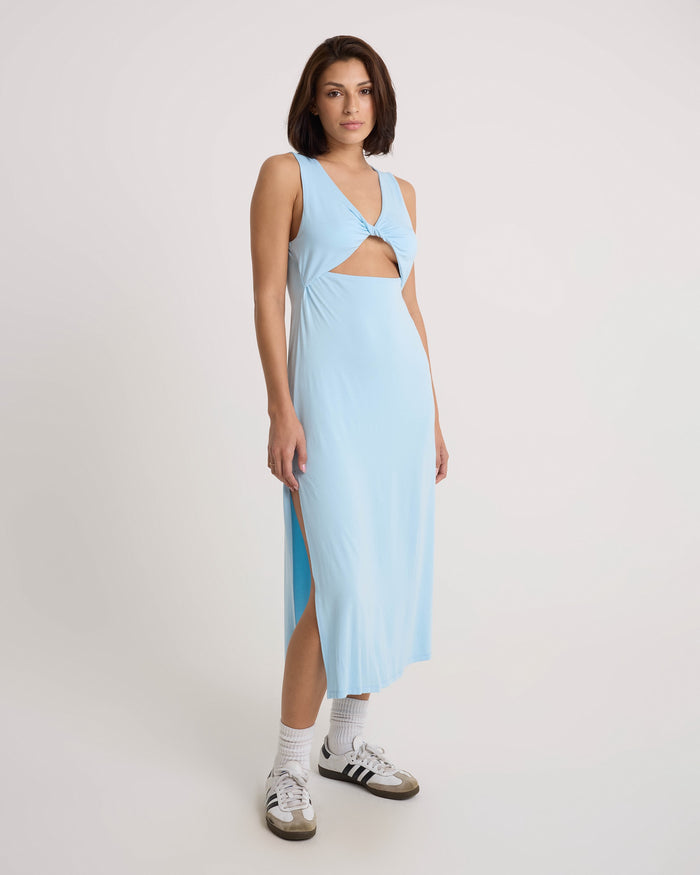 A woman stands against a plain backdrop wearing the Volcom Women's Stone Luz Dress, which features a sleeveless twist-front bodice and side slit. She pairs it with white socks and black Adidas sneakers.