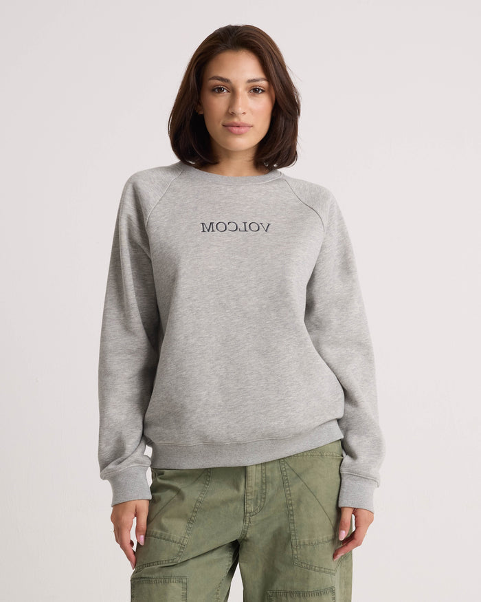 A woman stands against a plain light background with her hands by her sides, wearing the Volcom Women's Stone Magic BF pullover in heather gray and olive green pants. The relaxed fit of this sweatshirt adds to the laid-back vibe of her look.