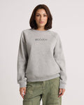 A woman stands against a plain light background with her hands by her sides, wearing the Volcom Women's Stone Magic BF pullover in heather gray and olive green pants. The relaxed fit of this sweatshirt adds to the laid-back vibe of her look.
