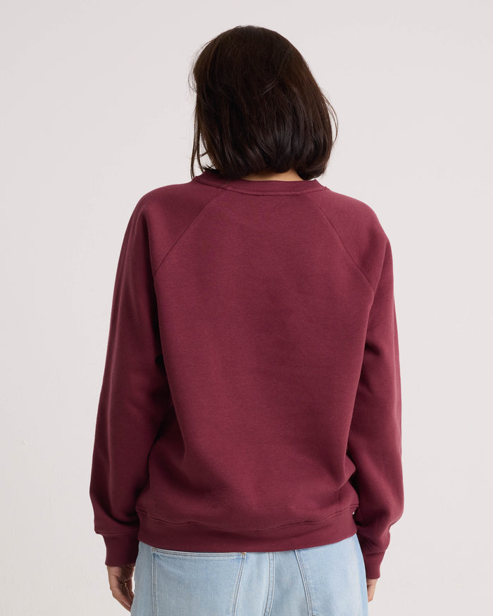 A person with shoulder-length dark hair is shown from the back against a plain light background, wearing the Volcom Women's Stone Magic BF pullover hoodie in maroon with a relaxed fit, paired with light blue jeans.