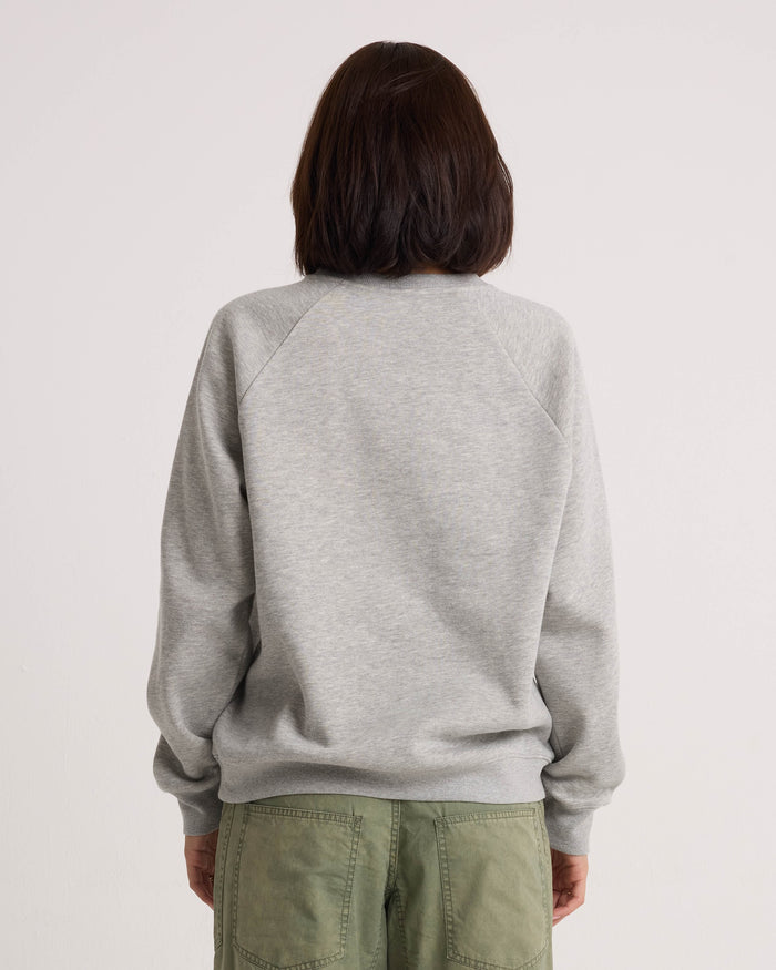 A person with shoulder-length dark hair, dressed in the Volcom Women's Stone Magic BF heather gray pullover hoodie and relaxed-fit olive green pants, stands facing away from the camera against a plain white background.
