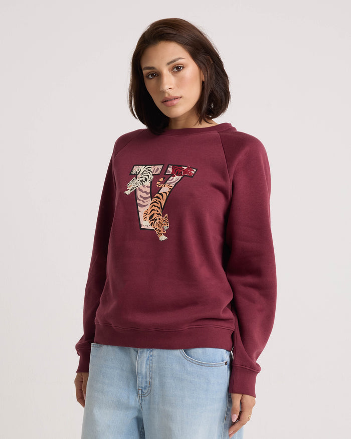 A woman wearing the Volcom Women's Stone Magic BF maroon relaxed fit sweatshirt, featuring a large V and tiger graphic on the front, paired with light blue jeans and standing against a plain background.