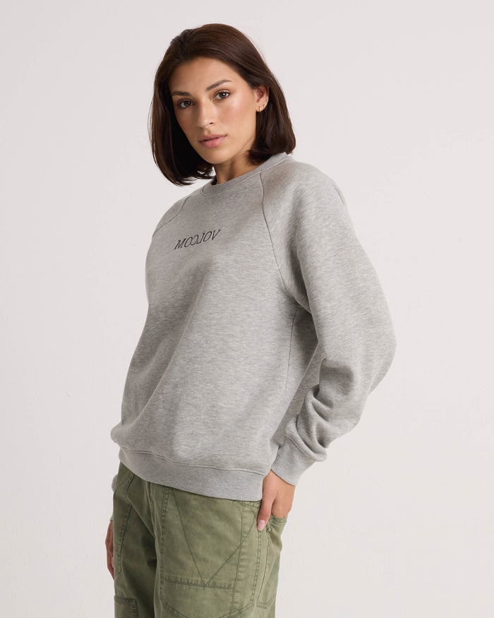 A woman stands against a plain background, wearing the Volcom Women's Stone Magic BF heather gray pullover hoodie with text on the chest and green cargo pants, looking at the camera with a neutral expression. The relaxed fit of her outfit from Volcom enhances the effortless vibe.