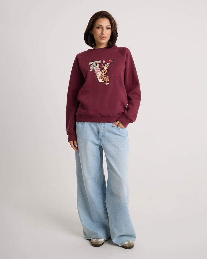 A woman wears the Volcom Women's Stone Magic BF maroon soft fleece pullover hoodie, which features a decorative letter V on the front. She pairs it with light blue wide-leg jeans and sneakers, standing against a plain light background. The relaxed fit of the hoodie brings effortless comfort to her casual look.