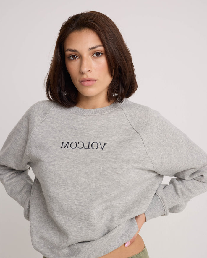 A woman with shoulder-length brown hair is wearing the Women's Stone Magic BF pullover hoodie by Volcom in a relaxed-fit heather gray style featuring the brand's logo printed in reverse, standing against a plain light background with a neutral expression.
