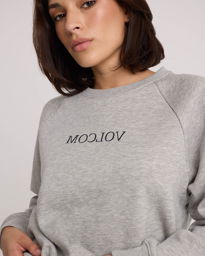 A person with shoulder-length brown hair wears the Volcom Women's Stone Magic BF sweatshirt in heather gray, featuring a relaxed fit and the word "VIOLCI" embroidered upside down in black letters on the front.