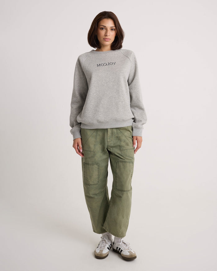 A woman with shoulder-length brown hair stands against a plain background, wearing the Volcom Women's Stone Magic BF heather gray pullover hoodie, relaxed fit green cargo pants, and white sneakers with black stripes.