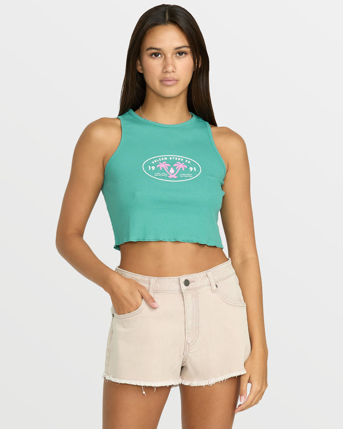 A woman wears the Volcom Women's Stone Mid Tank in green with a chest graphic, paired with light beige frayed-hem shorts, standing with one hand in her pocket against a plain light background.