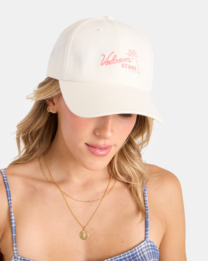 A woman in the Volcom Women’s Stone Rising Hat—ivory cotton with pink Volcom Stone text, a palm tree logo, and an adjustable strap—pairs it with a blue plaid top and layered gold necklaces, looking down against a light background.