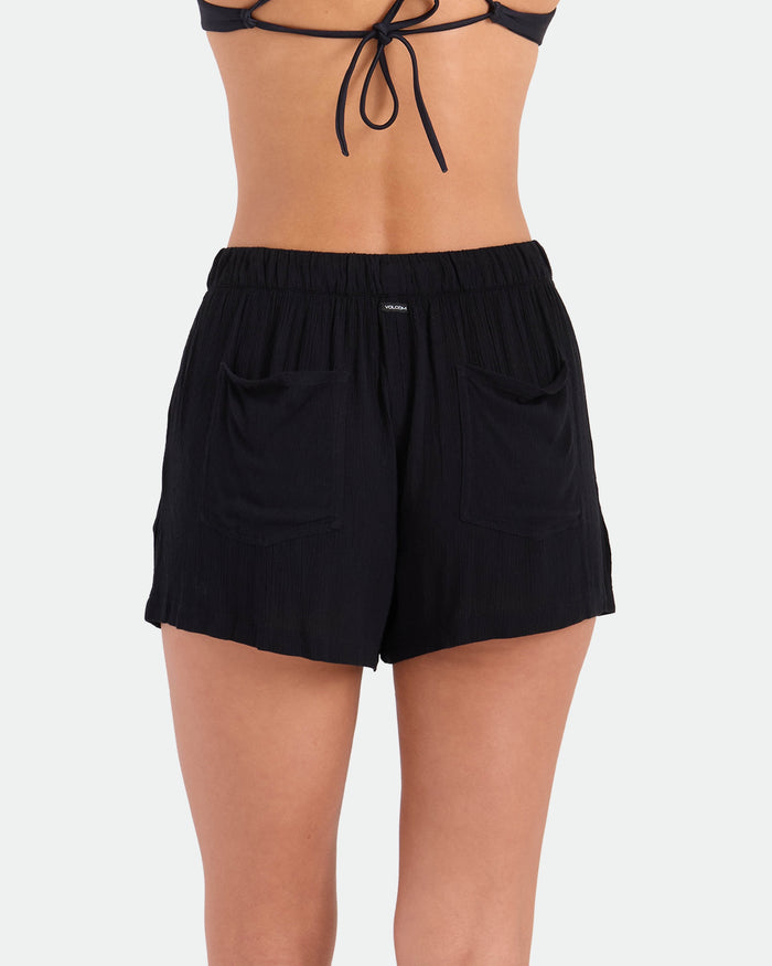 A woman is seen from behind in Volcom’s Women's Stone Shine Short Vsc, black viscose shorts with an elastic waistband and two back pockets, paired with a black string bikini top against a plain light background.