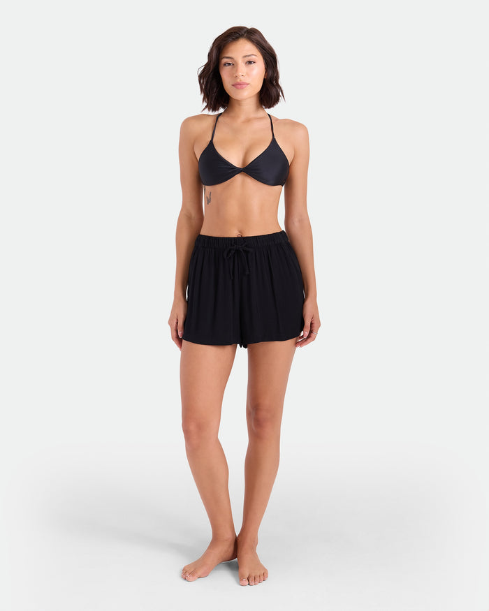 A woman stands barefoot against a white background, wearing a black bikini top and Volcom Women's Stone Shine Shorts Vsc with an elastic waistband. She faces forward, showing a neutral expression and relaxed posture.