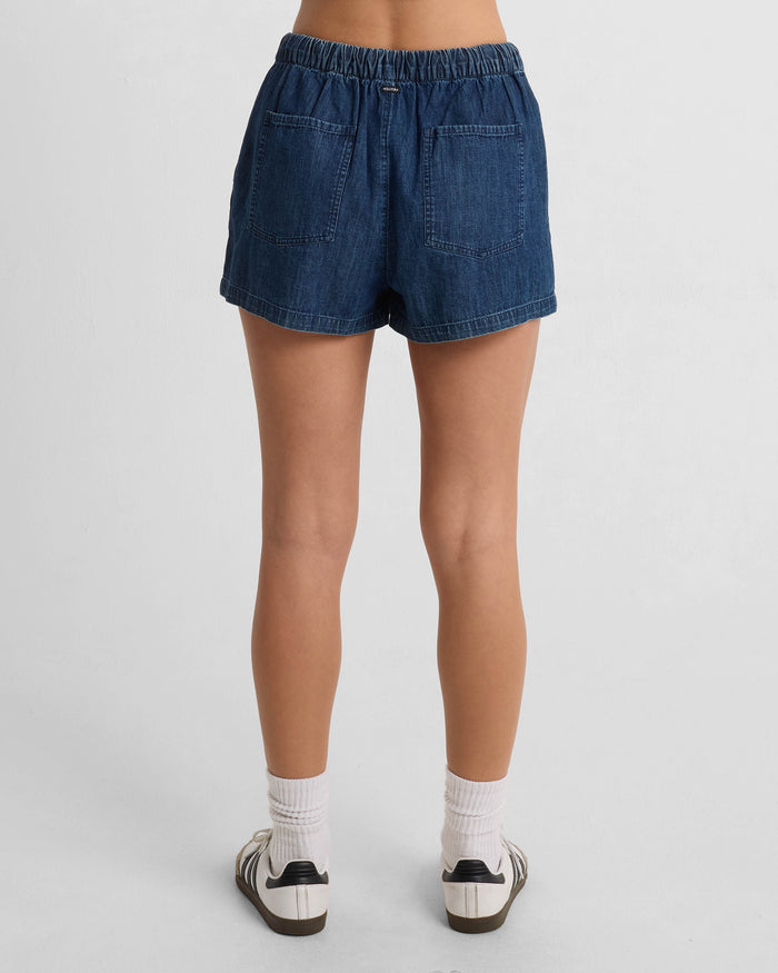 A person is standing facing away, wearing Volcom Women's Stone Shine Shorts made from 100% cotton with two back pockets, paired with white socks and white sneakers with black stripes, against a plain white background.