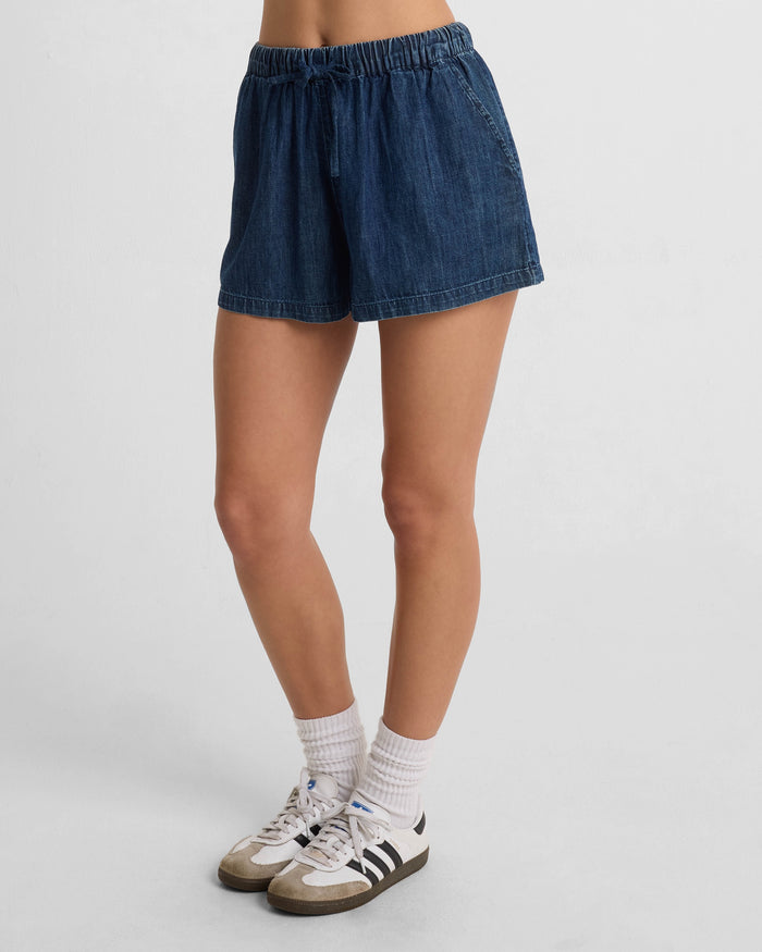 A person wears Volcom Women's Stone Shine Shorts with white socks and white sneakers featuring black stripes, standing against a plain white background.