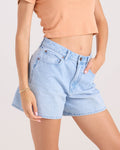 A person wearing Volcom Women's Stoned Bf Short in Blue Bird light wash (5-inch inseam, boyfriend fit) and a peach cropped tee, one hand in pocket. Only the midsection and upper legs show against a plain light background.