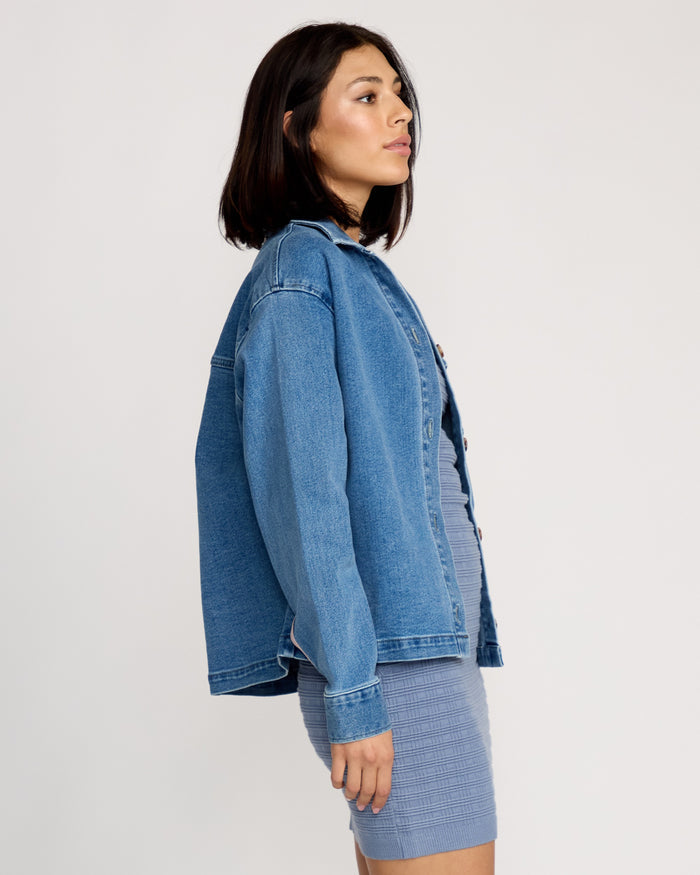 A woman with straight dark hair stands in profile, wearing the Volcom Women's Stoned BF Shacket over a fitted, textured light blue dress, against a plain white background.