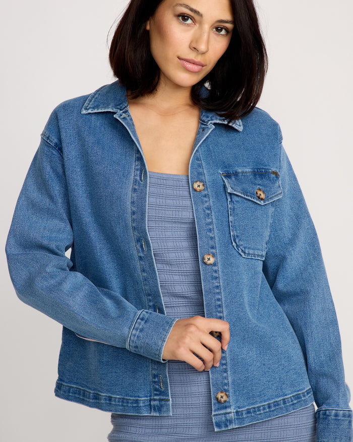 A woman with shoulder-length dark hair models the Volcom Women's Stoned BF Shacket over a textured, sleeveless light blue dress, standing against a plain, light-colored background.