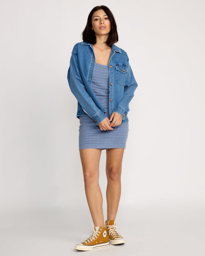 A woman with shoulder-length dark hair stands confidently against a plain backdrop wearing the Volcom Women's Stoned BF Shacket over a fitted blue dress and mustard yellow high-top sneakers.