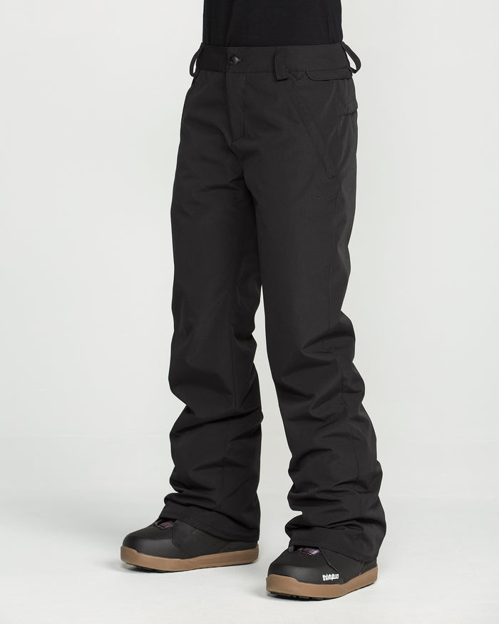 A person is wearing Volcom Women’s Stoned Insulated Pants in black along with black snow boots featuring brown soles, standing against a plain white background. Only the lower half of the body is visible.