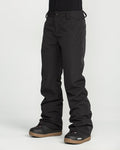A person is wearing Volcom Women’s Stoned Insulated Pants in black along with black snow boots featuring brown soles, standing against a plain white background. Only the lower half of the body is visible.