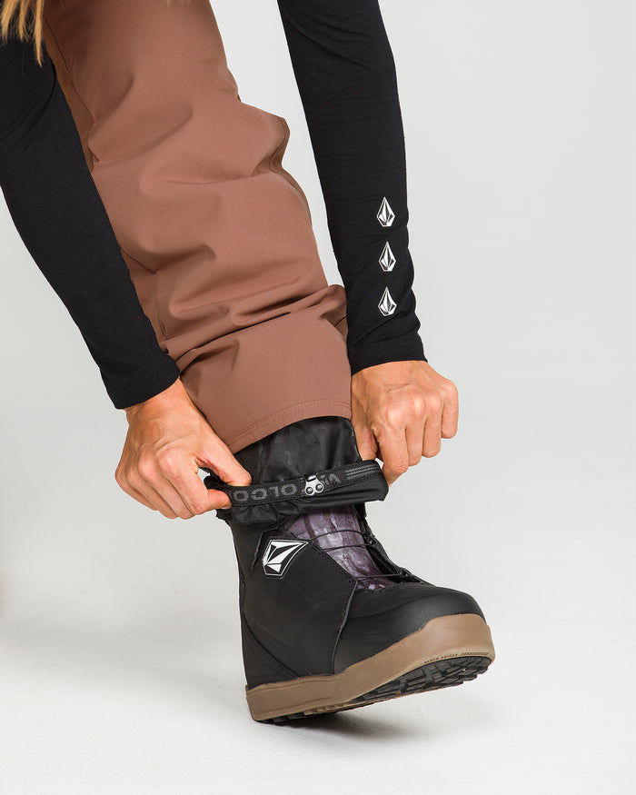 A person wearing Volcom Women’s Stoned Insulated Pants in brown—which offer recycled warmth and 10K waterproof protection—and a black long sleeve shirt tightens the Velcro strap on a black snowboarding boot.