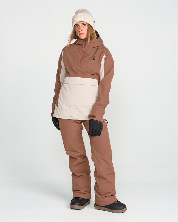 A person wearing a beige beanie, a brown and beige ski jacket, Volcom Women’s Stoned Insulated Pants for recycled warmth and 10K waterproof protection, black gloves, and black snow boots stands against a plain white background.