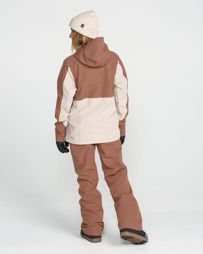 A person stands facing away against a plain white background, wearing the Volcom Women’s Stoned Insulated Pants featuring 10K waterproof protection and recycled insulation for sustainable warmth. They are also dressed in a brown and beige winter jacket, black gloves, a beige knit beanie, and snow boots.