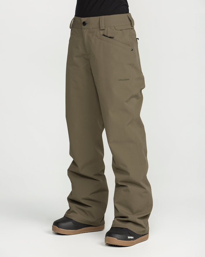 A person is shown from the waist down, standing against a plain white background, wearing Volcom Women’s Stoned Pants in olive green. The waterproof snow pants feature a relaxed fit and display a small logo on the left thigh. Black snow boots complete the look.