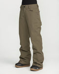 A person is shown from the waist down, standing against a plain white background, wearing Volcom Women’s Stoned Pants in olive green. The waterproof snow pants feature a relaxed fit and display a small logo on the left thigh. Black snow boots complete the look.